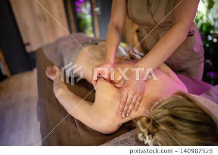 Relaxed woman client lying on spa bed in beauty salon receiving body treatment therapy from masseuse Relaxed woman client lying on spa bed in beauty salon receiving body treatment therapy from masseuse 140987526