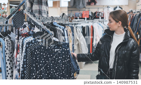 Fashionable shopper scanning clothing rack, comparing trendy outfits during retail shopping experience at modern boutique Fashionable shopper scanning clothing rack, comparing trendy outfits during retail shopping experience at modern boutique 140987531