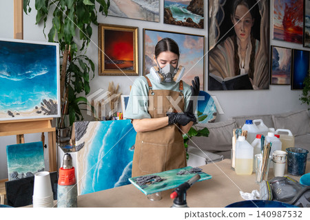 Serious woman artist preparing for work with epoxy resin, wearing gloves and respirator for safety. 140987532