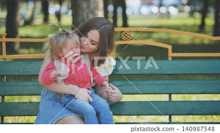 Compassionate mom soothing upset toddler daughter while seated on park bench during bright summer afternoon Compassionate mom soothing upset toddler daughter while seated on park bench during bright summer afternoon 140987548