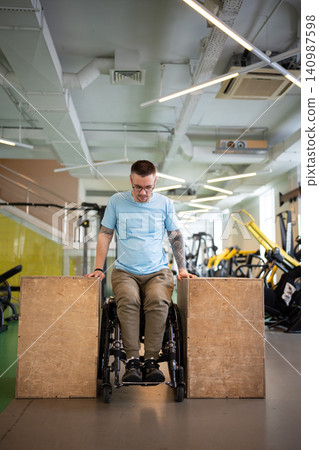 Confident man athlete in wheelchair doing push-ups strengthening exercise on wooden cubes in gym. Confident man athlete in wheelchair doing push-ups strengthening exercise on wooden cubes in gym. 140987598