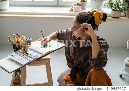 Talented woman spending free time with art hobby, painting pictures in sketchbook with aquarelle. Talented woman spending free time with art hobby, painting pictures in sketchbook with aquarelle. 140987617