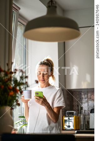 Relaxed thoughtful woman drinking coffee scrolling smartphone at cozy sunny kitchen beside window. Relaxed thoughtful woman drinking coffee scrolling smartphone at cozy sunny kitchen beside window. 140987646