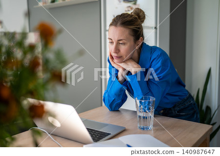 Thoughtful mature woman engaged in self learning process online on laptop at home in kitchen. Thoughtful mature woman engaged in self learning process online on laptop at home in kitchen. 140987647