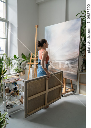 Talented focused woman painter artist working in home studio, looking at canvas painting on easel. 140987700