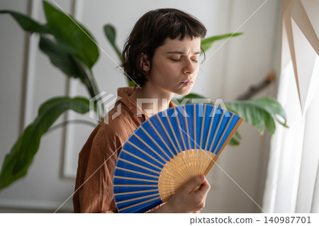 Woman at home annoyed by heatwave and heavy headache holding waving paper fan to cool down. Woman at home annoyed by heatwave and heavy headache holding waving paper fan to cool down. 140987701