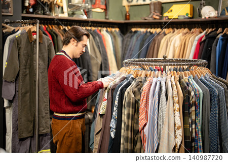 Handsome stylish man mindfully inspecting different shirts displayed on hanger in vintage shop. Handsome stylish man mindfully inspecting different shirts displayed on hanger in vintage shop. 140987720