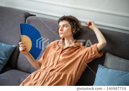 Tired woman lying resting on sofa waving hand paper fan, suffering from summer heat, annoyed face. Tired woman lying resting on sofa waving hand paper fan, suffering from summer heat, annoyed face. 140987739