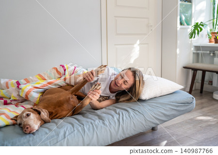 Laughing happy woman lying in bed with Vizsla dog, fooling playing around with pet on weekend. Laughing happy woman lying in bed with Vizsla dog, fooling playing around with pet on weekend. 140987766