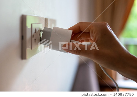 Woman hand holding mobile charger plugging in outlet on wall in room, problematic charging in hotels 140987790