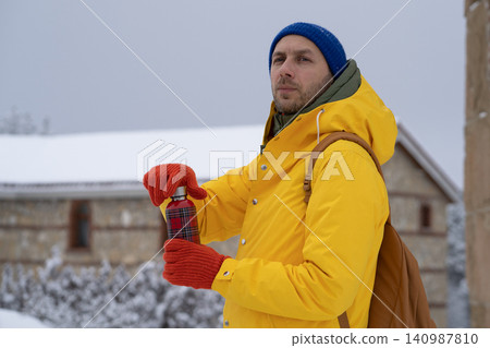 Thoughfull man in a yellow jacket holds thermos with hot drink in hands on snow winter day outdoors. Thoughfull man in a yellow jacket holds thermos with hot drink in hands on snow winter day outdoors. 140987810