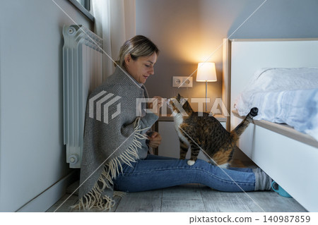 Young woman covered with warm blanket plays with cat feeding animal with food sitting on floor Young woman covered with warm blanket plays with cat feeding animal with food sitting on floor 140987859
