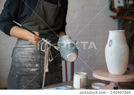 Female artisan in apron creates ceramic tableware painting white vase with brush in workshop 140987866