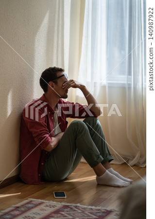 Upset depressed young man sitting on floor at home, received unexpected bad news over phone Upset depressed young man sitting on floor at home, received unexpected bad news over phone 140987922