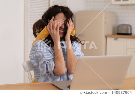 Mom work on laptop at home during lockdown, child distracts from work, covering her mother's eyes. Mom work on laptop at home during lockdown, child distracts from work, covering her mother's eyes. 140987964