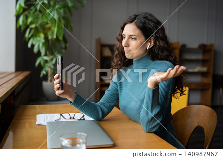 Smiling young businesswoman in earbuds sitting at office desk talking by video call on smartphone Smiling young businesswoman in earbuds sitting at office desk talking by video call on smartphone 140987967