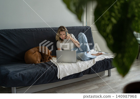 Woman lying on couch with her Vizsla dog, talking on cellphone, remotely working on computer laptop Woman lying on couch with her Vizsla dog, talking on cellphone, remotely working on computer laptop 140987968