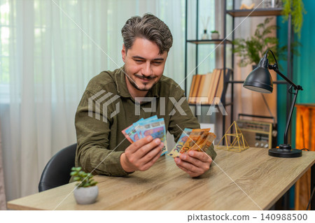 Middle-aged man counts cash smiling showing big financial success euro bills wealth at table desk Middle-aged man counts cash smiling showing big financial success euro bills wealth at table desk 140988500