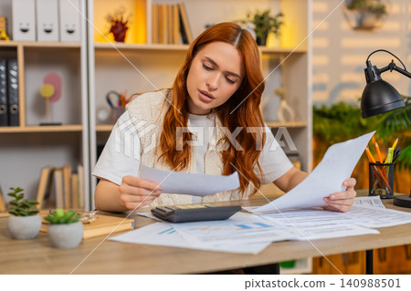 Young woman on home table paying bills with calculator and checking receipts during home budget task Young woman on home table paying bills with calculator and checking receipts during home budget task 140988501