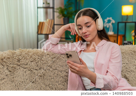 Young woman listening to podcast on sofa wearing headphones nodding while writing insightful notes Young woman listening to podcast on sofa wearing headphones nodding while writing insightful notes 140988503