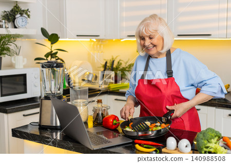 Senior old woman cooking vegetables in kitchen using laptop recipe for healthy diet home lifestyle Senior old woman cooking vegetables in kitchen using laptop recipe for healthy diet home lifestyle 140988508
