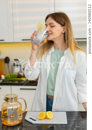 Middle-aged woman drinking lemon water in home kitchen for hydration wellness diet weight loss plan Middle-aged woman drinking lemon water in home kitchen for hydration wellness diet weight loss plan 140988512