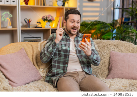 Young man on home sofa writing idea in notebook with raised finger and relieved smile after insight Young man on home sofa writing idea in notebook with raised finger and relieved smile after insight 140988533