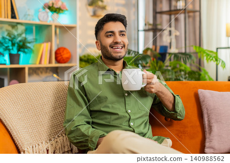 Indian man on home sofa enjoying hot drink and smiling peacefully during cozy quiet home relaxation Indian man on home sofa enjoying hot drink and smiling peacefully during cozy quiet home relaxation 140988562