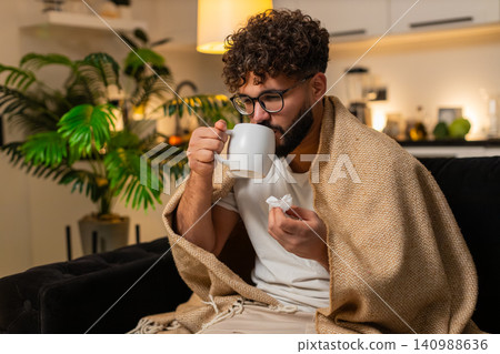 Arabian man sick with sore throat drinks hot tea with medicine uses paper tissues at night indoors 140988636
