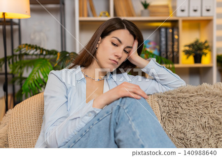 Young woman on home sofa feeling sad and worried thinking deeply and shaking head in concern indoors Young woman on home sofa feeling sad and worried thinking deeply and shaking head in concern indoors 140988640