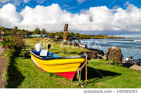 A vibrant yellow boat rests on green grass near the coast with a historic Moai statue standing in the background at Caleta Hanga Roa, Easter Island, Chile. A vibrant yellow boat rests on green grass near the coast with a historic Moai statue standing in the background at Caleta Hanga Roa, Easter Island, Chile. 140989265