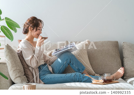 一位穿著時尚的亞洲女性坐在客廳的沙發上,在下午茶時間閱讀雜誌。 一位穿著時尚的亞洲女性坐在客廳的沙發上,在下午茶時間閱讀雜誌。 140992335