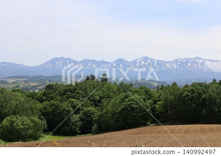 Hokkaido in June, Hokkaido in spring, Hokkaido in early summer: The town of Biei and the Tokachi Mountain Range viewed from the Northwest Hill Observation Park in Biei, a town of rolling hills. Hokkaido in June, Hokkaido in spring, Hokkaido in early summer: The town of Biei and the Tokachi Mountain Range viewed from the Northwest Hill Observation Park in Biei, a town of rolling hills. 140992497