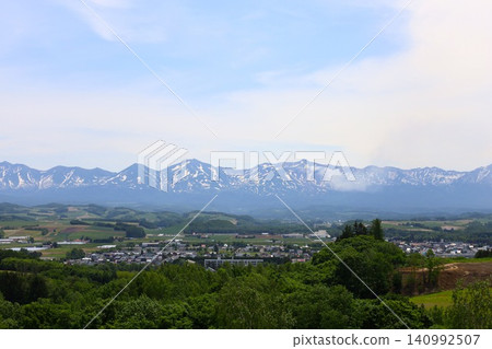 Hokkaido in June, Hokkaido in spring, Hokkaido in early summer: The town of Biei and the Tokachi Mountain Range viewed from the Northwest Hill Observation Park in Biei, a town of rolling hills. Hokkaido in June, Hokkaido in spring, Hokkaido in early summer: The town of Biei and the Tokachi Mountain Range viewed from the Northwest Hill Observation Park in Biei, a town of rolling hills. 140992507