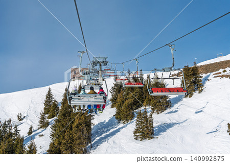 Skiers riding a chairlift over snowy slopes in Saalbach, Austria on a bright sunny winter day Skiers riding a chairlift over snowy slopes in Saalbach, Austria on a bright sunny winter day 140992875