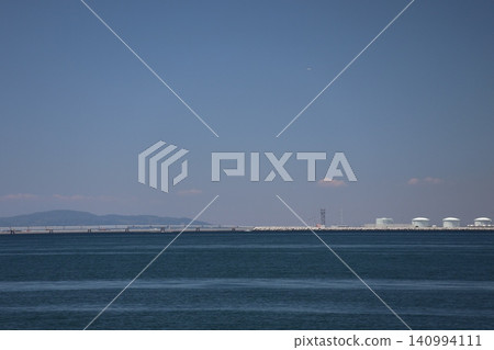 A seascape featuring a blue sky, clouds, mountains, an approach light bridge, fuel tanks, and the blue surface of the ocean. 140994111