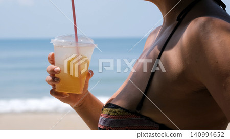 Close-up of young woman in swimwear holding a fresh iced lemonade drink in hand on a sunny tropical beach. Refreshment concept Close-up of young woman in swimwear holding a fresh iced lemonade drink in hand on a sunny tropical beach. Refreshment concept 140994652