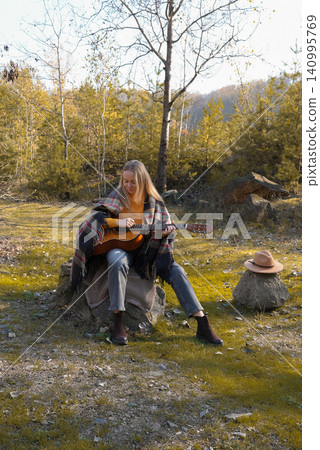 Beautiful blonde girl playing acoustic guitar and singing relaxing enjoying outdoors on sunny autumn day. Hippie woman in boho style learning and practicing playing musical instrument. Fall leaves Beautiful blonde girl playing acoustic guitar and singing relaxing enjoying outdoors on sunny autumn day. Hippie woman in boho style learning and practicing playing musical instrument. Fall leaves 140995769