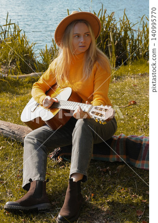 Portrait of smiling caucasian artist plays wooden acoustic guitar in autumnal park. Young songwriter plays string musical instrument hobby outside in nature fall time. Audio music healing Portrait of smiling caucasian artist plays wooden acoustic guitar in autumnal park. Young songwriter plays string musical instrument hobby outside in nature fall time. Audio music healing 140995770