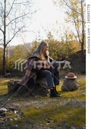 Blonde woman playing string guitar outdoors in autumn forest. Concept of sound therapy, mental health and wellness rituals. Calmness tranquility audio-sensory practices. Aura farming energy 140995776