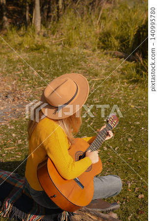 Beautiful blonde girl playing acoustic guitar and singing relaxing enjoying outdoors on sunny autumn day. Hippie woman in boho style learning and practicing playing musical instrument. Fall leaves Beautiful blonde girl playing acoustic guitar and singing relaxing enjoying outdoors on sunny autumn day. Hippie woman in boho style learning and practicing playing musical instrument. Fall leaves 140995780