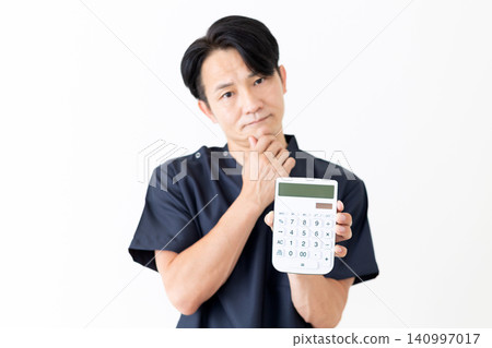 A middle-aged male doctor in navy blue scrubs ponders with a calculator in his hand. 140997017