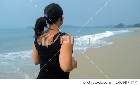 Back view of young woman in black sportswear with ponytail running along a sandy coastal beach with sea waves and blue sky. Beach running concept Back view of young woman in black sportswear with ponytail running along a sandy coastal beach with sea waves and blue sky. Beach running concept 140997477