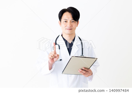 A middle-aged male doctor in a white coat holds a binder of medical charts and raises his index finger. A middle-aged male doctor in a white coat holds a binder of medical charts and raises his index finger. 140997484
