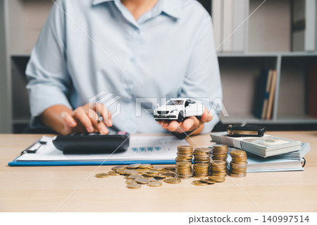 Person calculating auto financing while holding a white toy car, with calculator, cash, and coins on desk, representing car loan budgeting, vehicle purchase cost, and finance management. 140997514