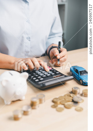 Business person calculating auto loan budget with piggy bank, blue toy car, coins, and calculator, representing vehicle finance, car insurance cost, and purchase planning. 140997517