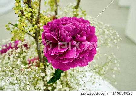 A vibrant pink peony flower surrounded by baby's breath and green berries. A vibrant pink peony flower surrounded by baby's breath and green berries. 140998026