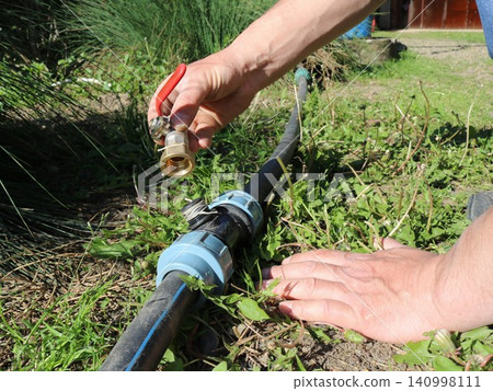 The hands of a technician attaching a brass ball valve with a red handle to a plastic tee fitting on a black HDPE pipe. The irrigation system is being installed on the grass at a summer cottage. The hands of a technician attaching a brass ball valve with a red handle to a plastic tee fitting on a black HDPE pipe. The irrigation system is being installed on the grass at a summer cottage. 140998111