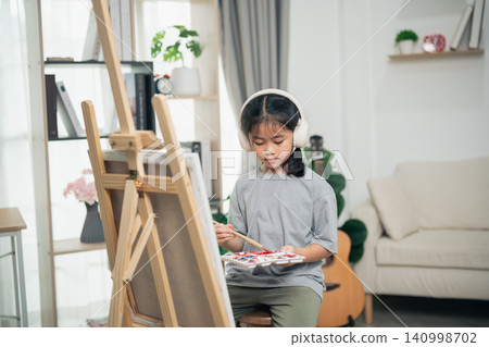 Young girl wearing headphones painting on canvas using brush and palette in cozy bright living room with artistic decor and comfortable furniture Young girl wearing headphones painting on canvas using brush and palette in cozy bright living room with artistic decor and comfortable furniture 140998702