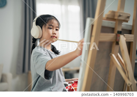 Young Asian girl wearing white headphones painting on easel with focused expression in cozy indoor room with soft natural light 140998703
